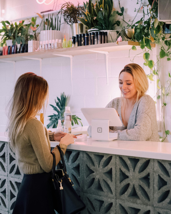 A photograph of a boutique restaurant. Guest interacting with a smiling host at the cash register.